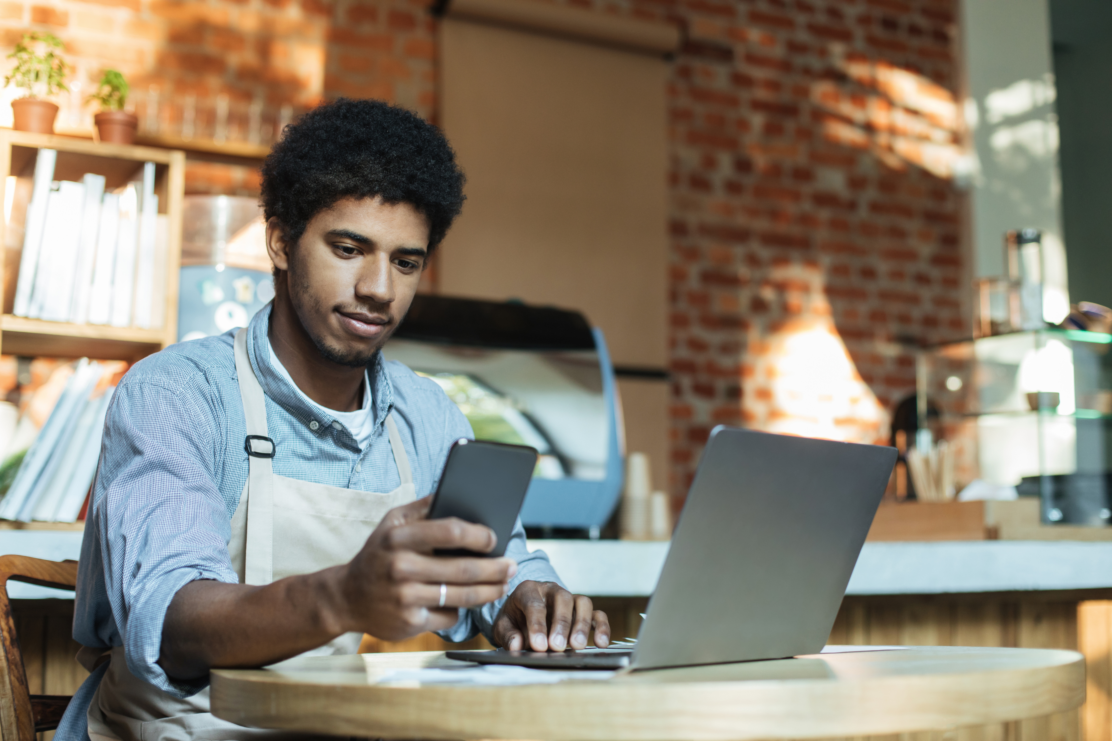 Man in apron sits at table and works in smartphone and laptop in interior of loft coffee shop